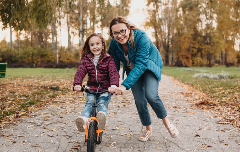 Caucasian mother looking through her eyeglasses is helping her daughter to ride the bike during an autumn walk
