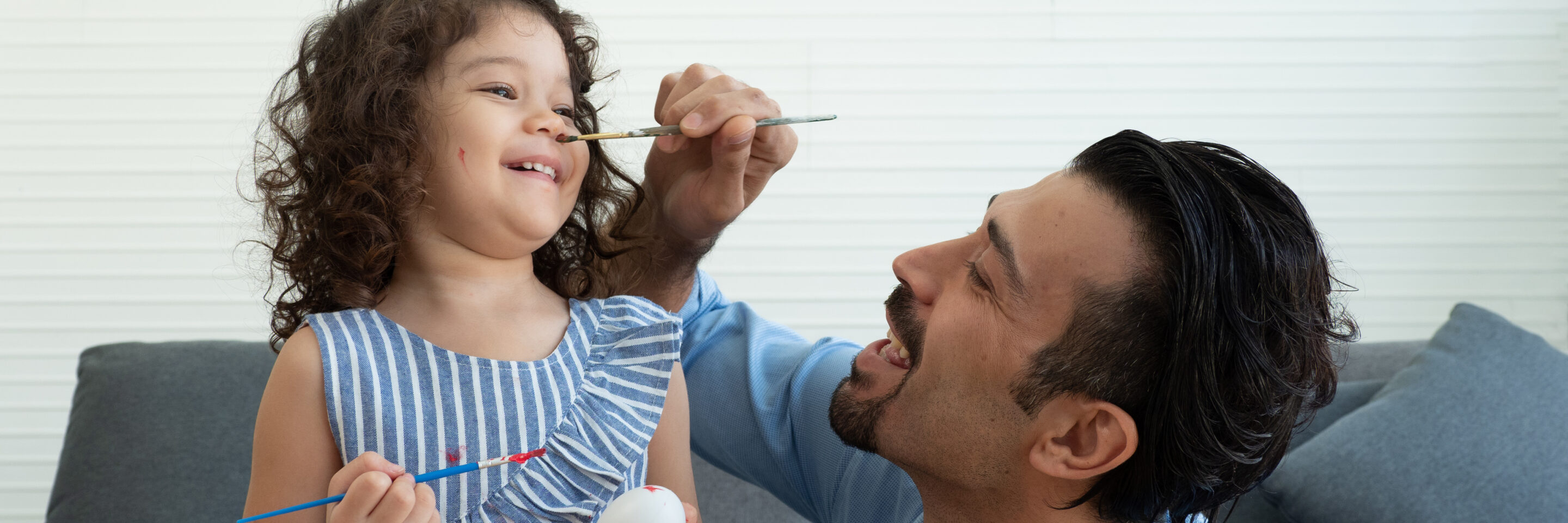 Father trying to paint on nose of cute little toddler daughter while coloring the Easter Eggs together. Happy Easter concept.