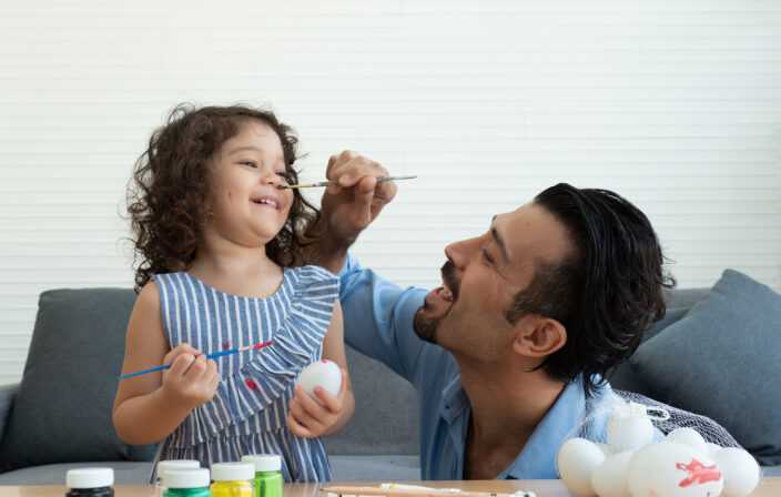 Father trying to paint on nose of cute little toddler daughter while coloring the Easter Eggs together. Happy Easter concept.